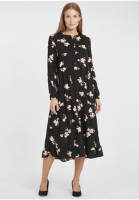 Zwarte jurk met lange mouwen en een knoopsluiting aan de voorkant, met een bloemenpatroon in roze en wit. De rok loopt uit met een gerimpelde zoom.