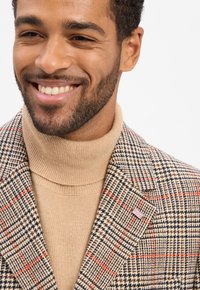 Lächelnder Mann mit kurzen Locken trägt einen beigen Rollkragenpullover und ein braunes gemustertes Sakko mit einer kleinen Union-Jack-Nadel am Revers.