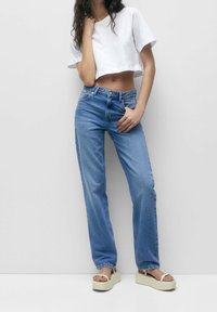 Camiseta blanca cortada, jeans de tiro alto azules y sandalias de plataforma beige. Tela suave con un ajuste relajado; textura de mezclilla visible.