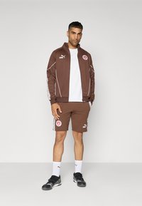 Chándal deportivo marrón con acentos blancos, que incluye una chaqueta con cremallera y pantalones cortos a juego. Incluye logotipos de la marca y patrones laterales distintivos.