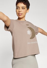 Světle hnědé bavlněné tričko s krátkými rukávy, které zobrazuje grafiku měsíce a text "YOGA SEARCHER" v bílé barvě. Zkrácený design.