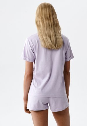 Kobieta z długimi blond włosami, ubrana w jasnofioletową bluzkę z krótkim rękawem i pasujące szorty, stojąca tyłem na jednolitym tle.
