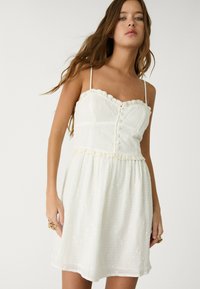 Vit bomullssundress med spetskant, spaghetti-remmar och knappdetalj. Lätt texturerat tyg med blommig broderiaccent.