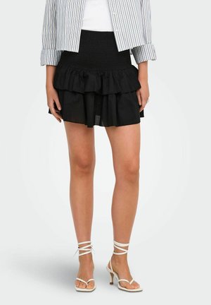 Kobieta w czarnej, warstwowej mini spódnicy, białych sandałkach na obcasie z paskami oraz niebiesko-białej koszuli w paski z podwiniętymi rękawami.