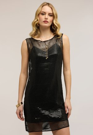 Blonde Frau in einem ärmellosen schwarzen Paillettenkleid mit goldenen Schmuckstücken, die vor einem schlichten hellen Hintergrund steht.