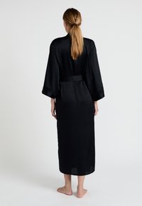 Robe en satin noir avec une coupe longue et fluide, des manches larges et une ceinture nouée à la taille, mettant en avant des accents de dentelle discrète aux bords des manches.