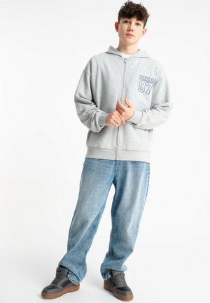 Nastolatek ubrany w jasnoszarą bluzę z kapturem na zamek z napisem "BRKLN 97", luźne niebieskie dżinsy i czarne trampki, stoi z rękami splecionymi na białym tle.