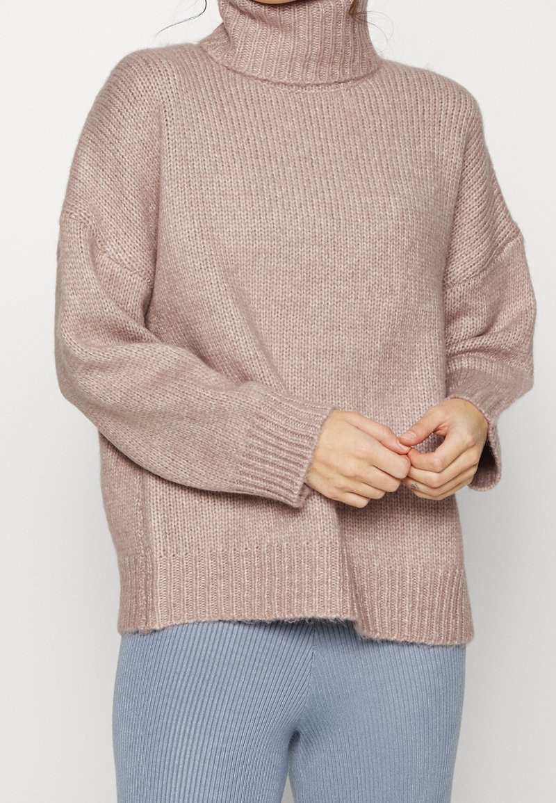 Femme portant un pull à col roulé en maille épaisse rose clair et un pantalon côtelé bleu clair, les mains jointes légèrement devant elle.