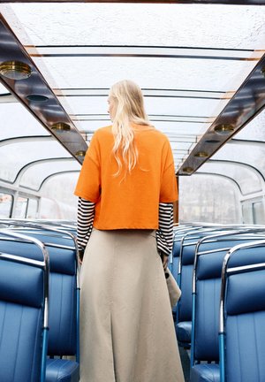 Persoon met lang blond haar, gekleed in een oranje top en een beige rok, staand in een voertuig met glazen dak en blauwe stoelen.