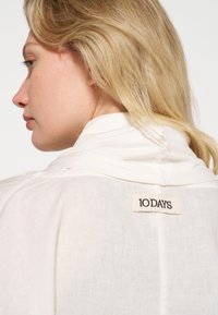 Witte linnen shirt met een kraag, voorzien van een geweven label op de achterkant met "10 DAYS" in zwart. Zachte textuur en lichte uitstraling.