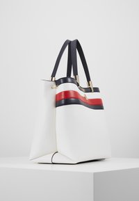 Valkoinen nahkainen kassi, jossa on tummansinisiä ja punaisia raitoja, kaksoiskahvat ja jämäkkä muoto, jossa on kultaisia yksityiskohtia.