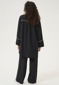 Melnā vieglā tunika ar garām piedurknēm, kurai ir krēmkrāsas apdare gar pleciem un piedurknēm, apvienota ar brīvām melnām biksēm.