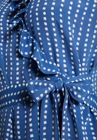 Vestido azul estampado con lunares blancos y detalle de volantes en el escote. Un lazo atado en la cintura realza el diseño.