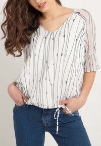 Blouse ample à manches courtes en blanc et marine, présentant des rayures verticales et une taille cintrée avec un lien. Manches transparentes ornées de volants.