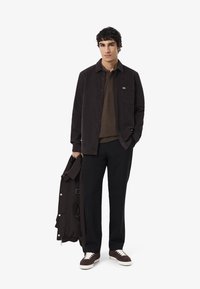 Camicia scura, con bottoni, sopra una polo marrone, abbinata a pantaloni neri. Giacca marrone appoggiata sopra un braccio, con bottoni a pressione. Sneakers eleganti con suola bianca.