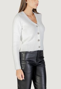 Cardigan bianco peloso con scollatura a V, dotato di cinque bottoni in strass, abbinato a pantaloni in pelle nera. Il cardigan ha una texture a coste.