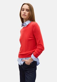 Maglione rosso realizzato in un morbido tessuto con scollo rotondo, abbinato a una camicia azzurro chiaro con bottoni sotto. I pantaloni sono jeans scuri.