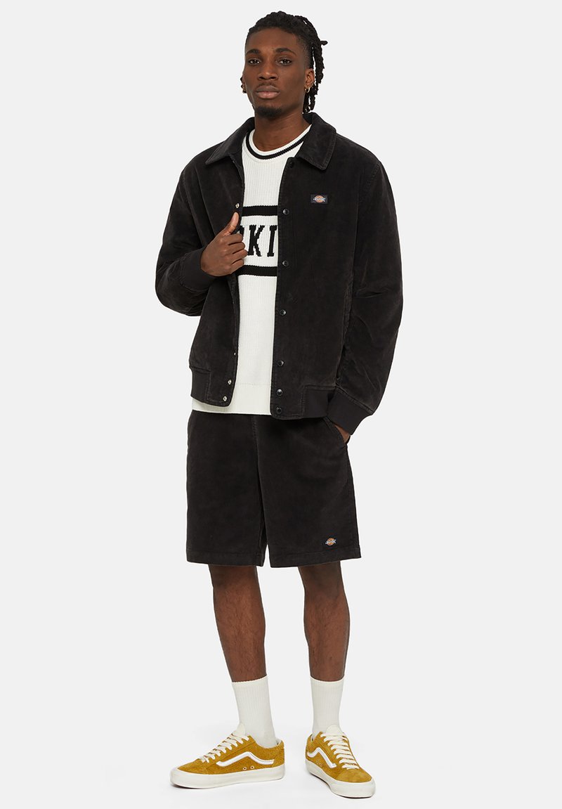 Veste en velours côtelé noire avec des boutons-pression, pull en tricot blanc avec du texte, short noir et baskets en daim jaunes avec des semelles blanches.