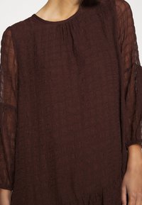 Kvinna iklädd en genomskinlig, mörkbrun strukturblus med långa ärmar och subtilt rutmönster.