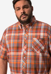 Košile s krátkým rukávem v kostičkovaném vzoru v oranžové, modré a bílé barvě. Obsahuje límec na knoflíček, přední kapsu a různé barevné vzory po celém povrchu.