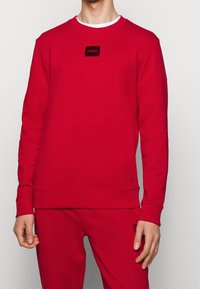 Homme portant un sweat-shirt rouge à manches longues et un pantalon rouge assorti avec des patchs logo noirs "HUGO" sur la poitrine et la cuisse.