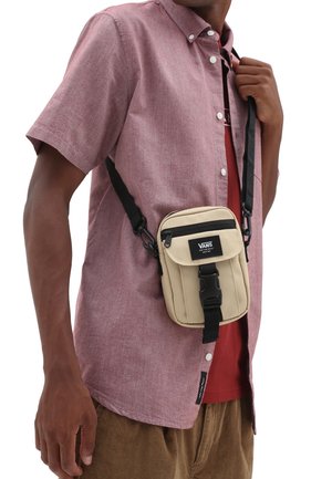 Personne portant une chemise rose à manches courtes avec boutons et un sac bandoulière Vans beige avec une sangle et une boucle noires, sur un t-shirt rouge.