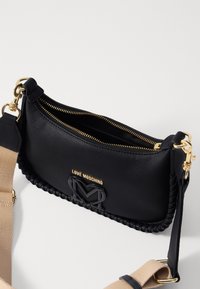 Černá kožená kabelka s texturou, opleteným okrajem, zlatým zipem a černým logem. Má odnímatelný béžový popruh.