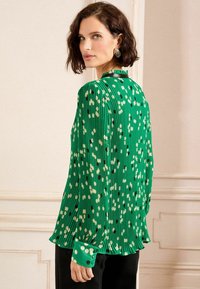 Blouse plissée verte avec des pois blancs et noirs, dotée de manches longues et d'un poignet à bouton. Texture lisse avec une coupe décontractée.