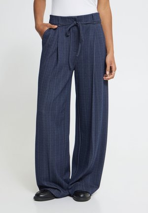 Pantalon large bleu marine à fines rayures avec ceinture nouée, porté avec des chaussures noires et un top blanc, une main dans la poche et l'autre détendue.