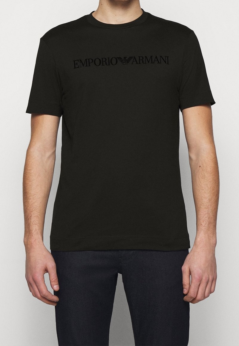 Homme portant un t-shirt noir à manches courtes Emporio Armani et un pantalon sombre, debout devant un fond clair uni.