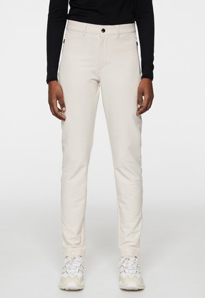 Beige, schmal geschnittene Hosen aus einem glatten, dehnbaren Material, mit einem vorderen Knopfverschluss und seitlichen Taschen. Getragen mit weißen Sneakers.