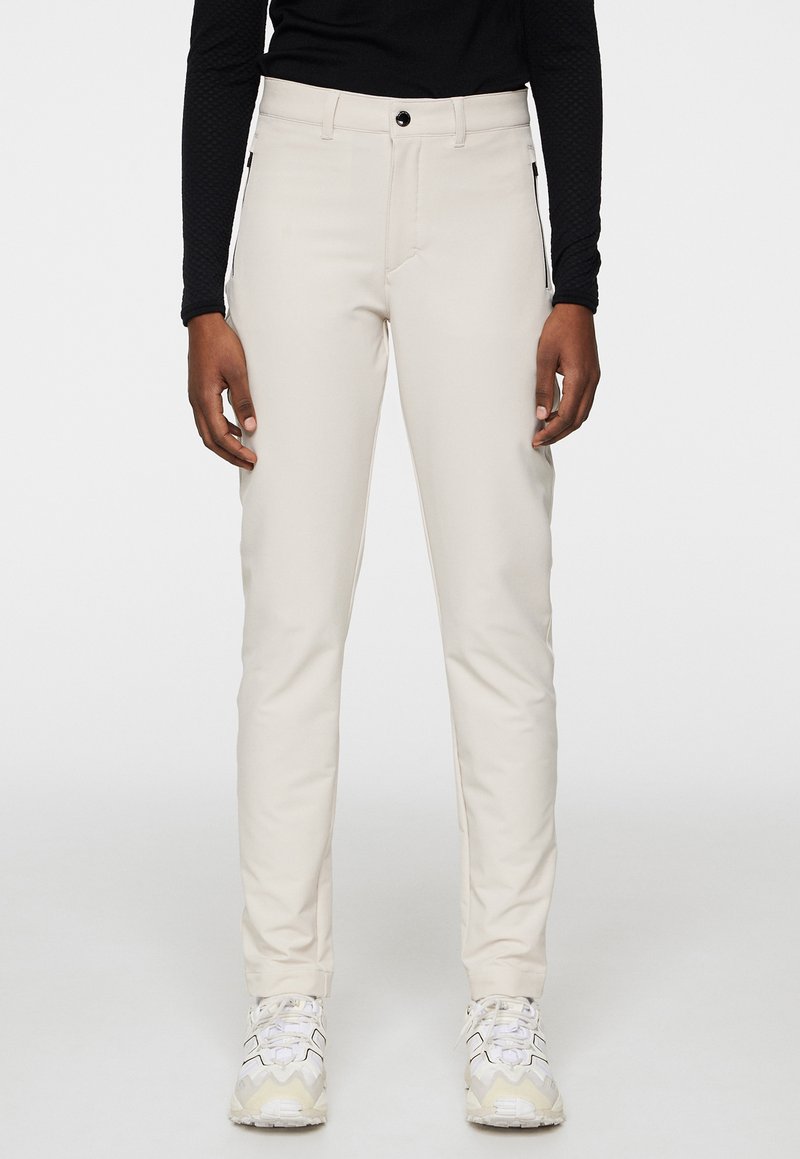 Beige, slim-fit broek gemaakt van een gladde, elastische stof, met een knoopsluiting aan de voorkant en zijzakken. Draagt met witte sneakers.