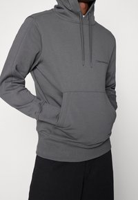 Šedá bavlněná mikina s kapucí, má přední klokaní kapsu, nastavitelnou kapuci se šňůrkou a decentní logo na hrudi.