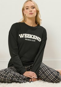 La maglietta nera a maniche lunghe presenta la scritta "WEEKEND BRUNCH CLUB" in morbido testo bianco, abbinata a pantaloni pigiama a quadri. Tessuto morbido e vestibilità rilassata.