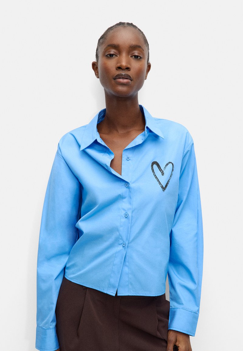 Jasnoniebieska koszula zapinana na guziki z długim rękawem, z czarnym konturem serca wyhaftowanym na lewej piersi. Gładkie wykończenie materiału.