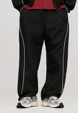 Personne portant un pantalon noir avec des bandes blanches sur les côtés, un t-shirt rouge, une veste noire et des baskets argentées et blanches, debout les mains dans les poches.