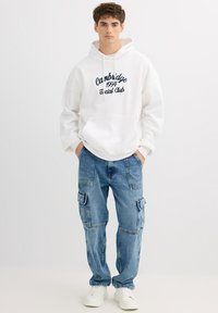 Sudadera blanca con bordado azul marino que dice "Cambridge 1994 Social Club", combinada con vaqueros cargo azul claro y zapatillas blancas.