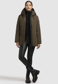 Young woman wearing an olive-green hooded jacket, black turtleneck, black leather pants, and black lace-up boots, standing with legs crossed.