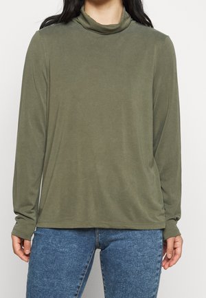 Femme portant un haut à col roulé vert olive à manches longues et un jean bleu, debout devant un fond clair uni.