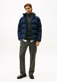 Doudoune bleu marine avec un design matelassé, portée sur un pull en charbon avec un motif en losange, associée à un pantalon large gris et des chaussures noires.