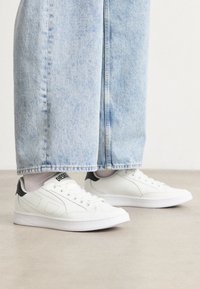 Vita sneakers med svart hälaccent, perforerat mönster och platt vit sula, kombinerade med ljusblå denimbyxor som är något kortade.