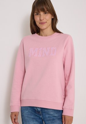 Femme portant un sweat-shirt rose clair avec le mot "MIND" brodé à l'avant, associée à un jean bleu, debout devant un fond uni.