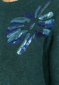 Teal trui met een fuzzy textuur, versierd met een bloemenpatroon van blauwe en turquoise pailletten. De pailletten zijn van verschillende grootte en zijn op verschillende plaatsen aangebracht.