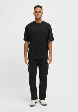 Ung mann stående, iført en svart oversized T-skjorte, svarte jeans og hvite sneakers med svarte detaljer, mot en ensfarget lys bakgrunn.