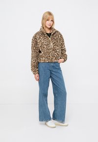 Kobieta w brązowej polarowej kurtce w cętki, niebieskich szerokich dżinsach i białych sneakersach, stojąca z jedną ręką w kieszeni na białym tle.