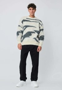 Pull en maille crème avec motifs abstraits noirs, col rond, épaules décalées, porté sur un pantalon noir et des baskets blanches.