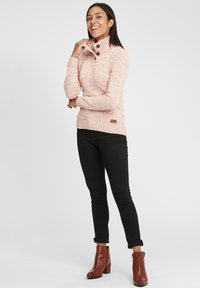 Maglione lavorato a maglia rosa chiaro con colletto alto e dettagli con bottoni. Polsini e orlo a costine. Pantaloni neri aderenti e stivaletti marroni alla caviglia.