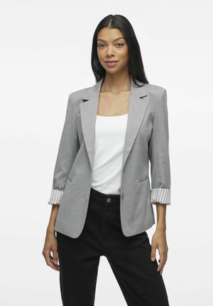 Blazer grigio con una vestibilità su misura, caratterizzato da polsini a righe e tasche laterali. Indossato sopra un top bianco e abbinato a pantaloni neri.