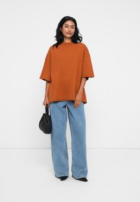 Υπέρμετρα γκρι τ-shirt σε καφέ απόχρωση, με κοντά μανίκια, συνδυασμένο με ανοιχτό μπλε παντελόνι σε φαρδιά γραμμή. Συμπληρωμένο με μια μικρή μαύρη τσάντα.