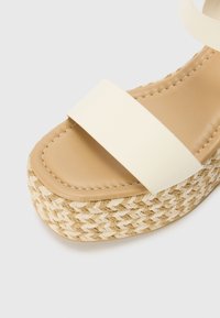 Sandale espadrille beige avec une lanière crème, dotée d'une semelle en jute tressé et d'une semelle intérieure lisse et rembourrée pour un confort optimal. Design minimaliste.